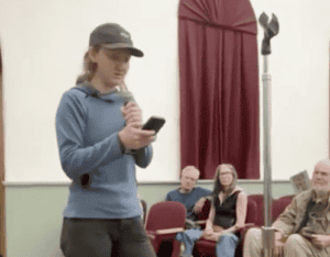 Person speaking at a community event, holding a microphone and phone, with an attentive audience seated in the background.
